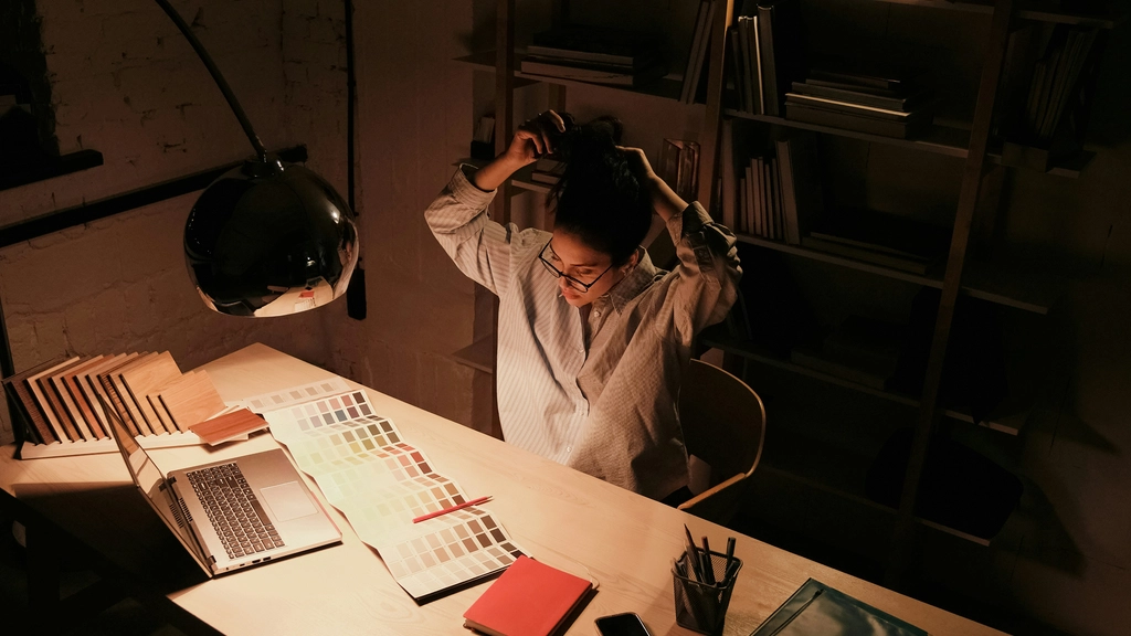 A person sits at their desk illuminated by a desk lamp with their hands behind their head