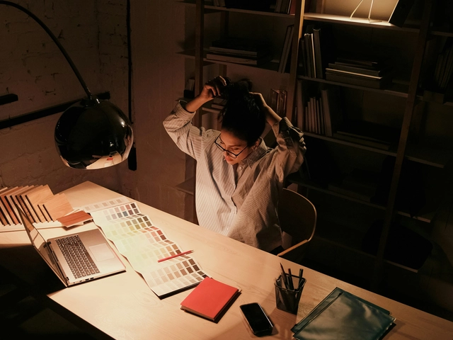 A person sits at their desk illuminated by a desk lamp with their hands behind their head