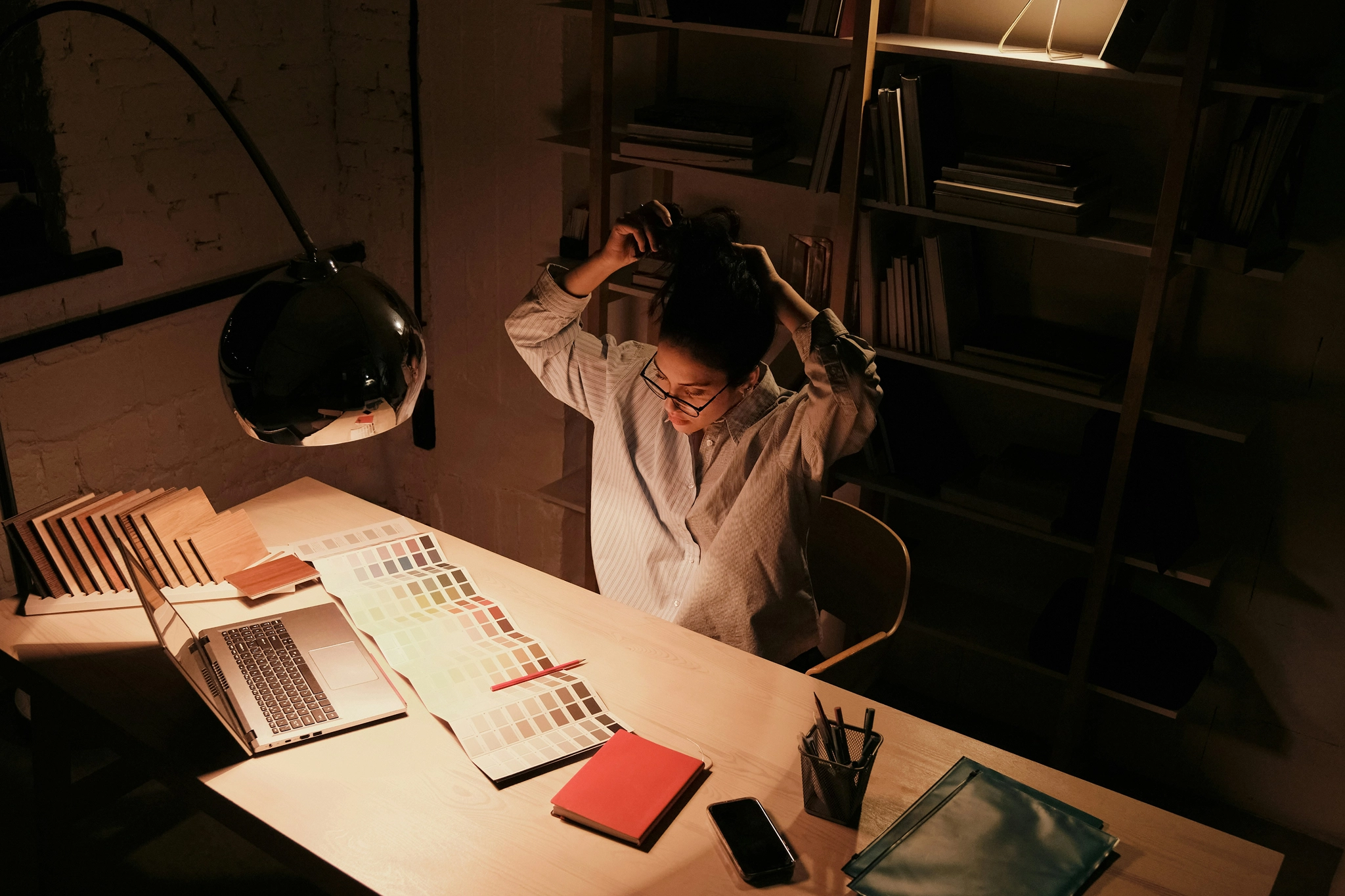 A person sits at their desk illuminated by a desk lamp with their hands behind their head