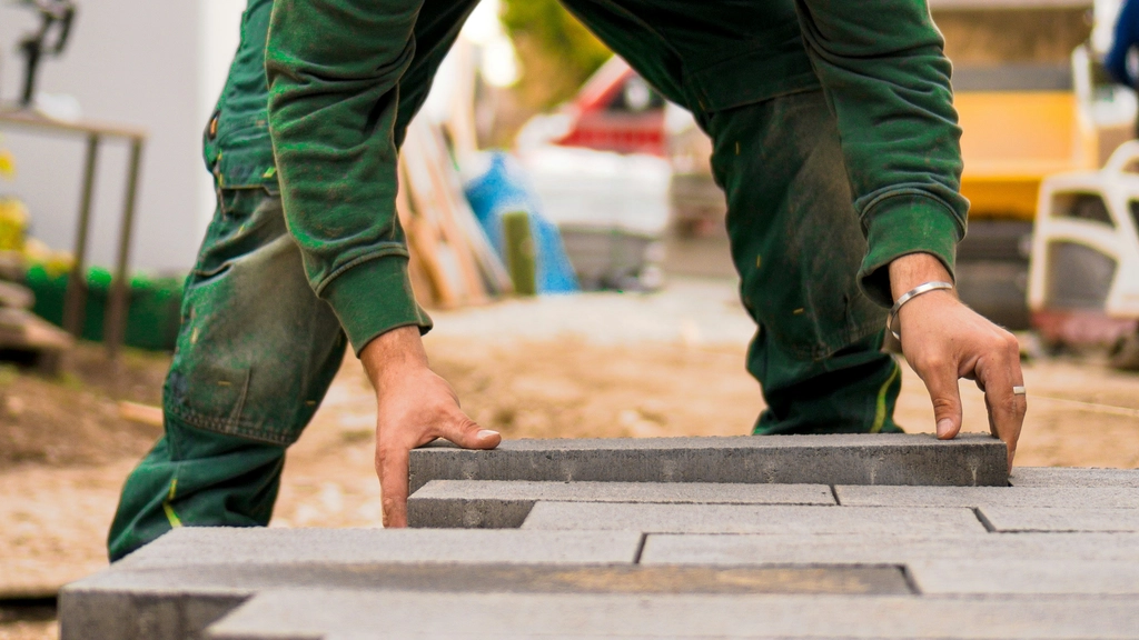 A person in a green shirt lays a brick on the ground, building a floor