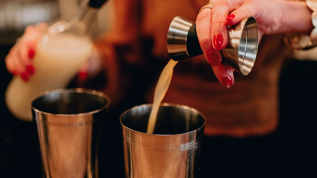 A person with long hair and long red nails pours a shot of liquid into a mixer behind a bar