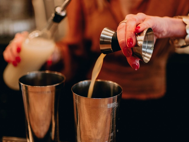 A person with long hair and long red nails pours a shot of liquid into a mixer behind a bar