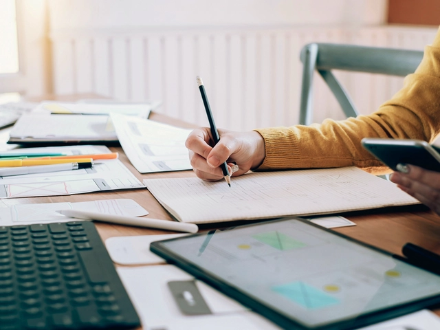 Hand writing on notebook on a desk filled with paperwork and a keyboard