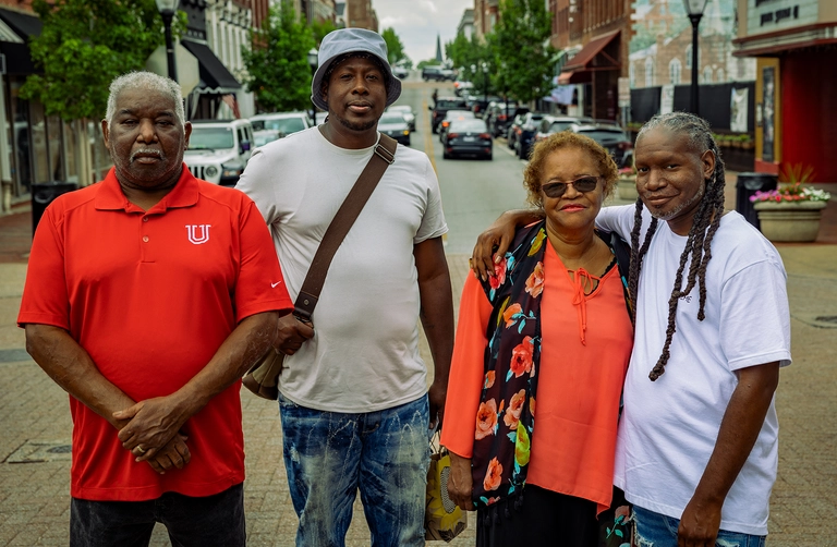 Group of four people standing together on a downtown street, posing with shops and parked cars in the background.