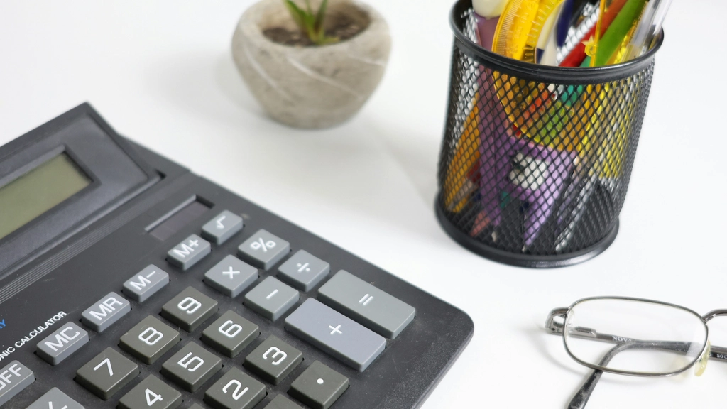 A calculator, small succulent plant a pen cup and glasses are all visible on a white desk 