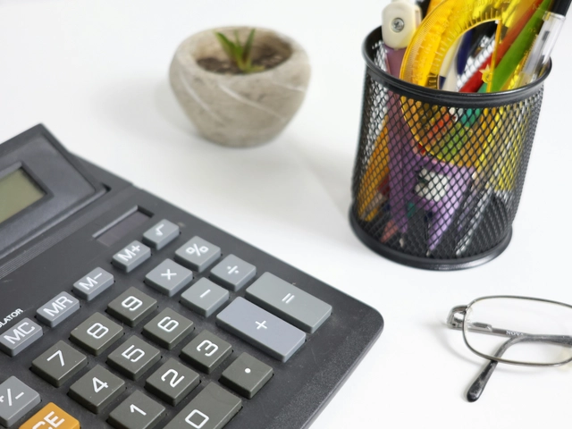 A calculator, small succulent plant a pen cup and glasses are all visible on a white desk 
