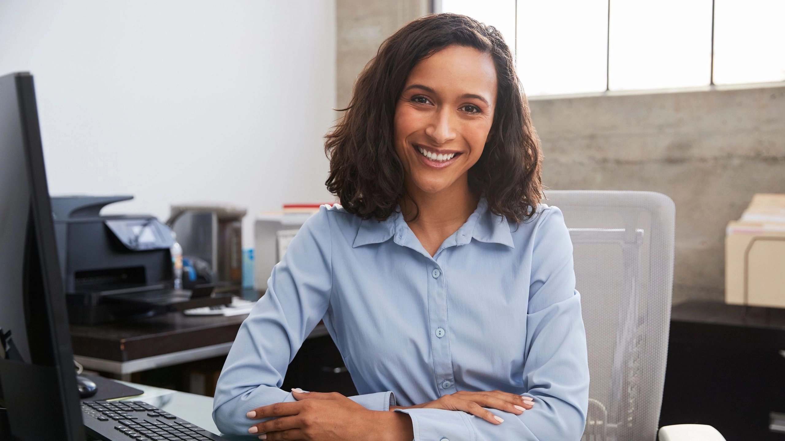 Woman with shoulder length brown hair wearing light blue shirt sitting at a desk and smiling at the camera to illustrate article about personal credit for business owners