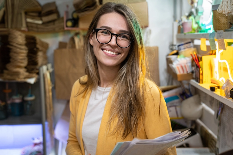 A female business owner with long hair and glasses in a yellow sweater holding paperwork and smiling to illustrate paying California franchise tax