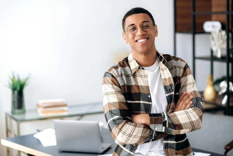 Successful confident young man with glasses, stylishly dressed, standing near work desk with arms crossed, looking at camera with friendly smile to illustrate article about business life stages.