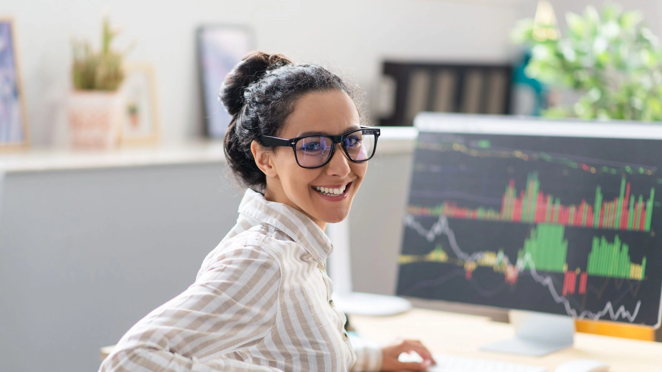 Woman with hair in bun and glasses sitting at desk in front of computer displaying charts and smiling to illustrate article about good profit margins
