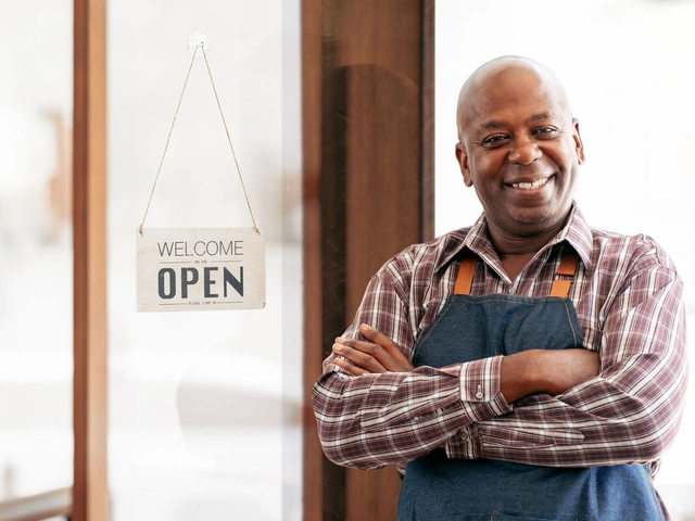 Man wearing Apron and checkered short smiling and standing in front of door with open sign to illustrate article about Small Business Saturday.