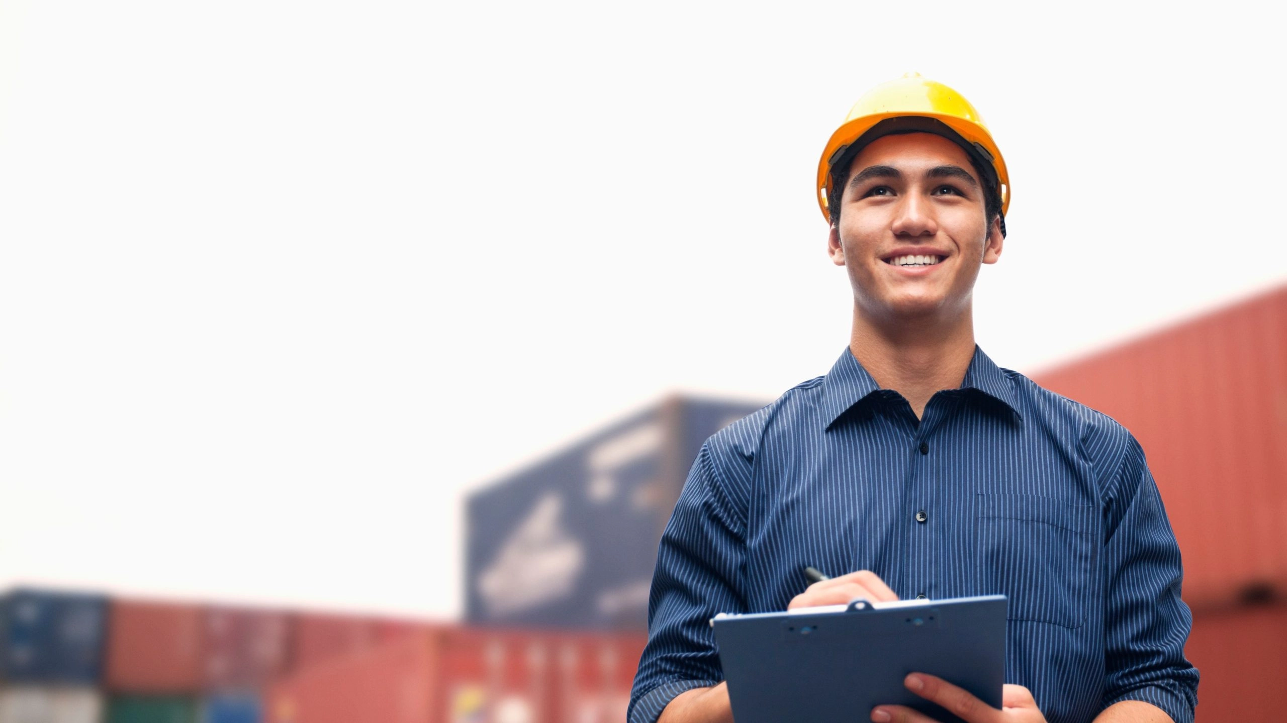 Smiling man wearing a yellow hard hat and blue shirt and holding a clipboard, standing in front of shipping containers to illustrate article about tariffs and small business owners.