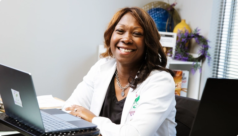 Smiling woman in a white coat sitting at a desk with a laptop