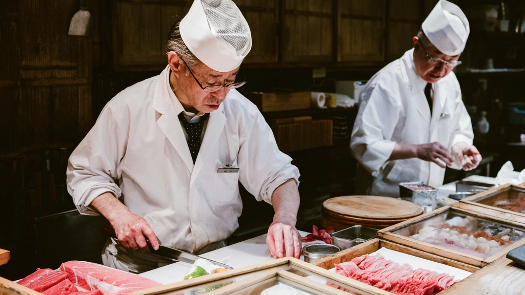 Two sushi chefs in traditional white uniforms preparing fresh sashimi behind a counter filled with trays of raw fish in a Japanese restaurant kitchen