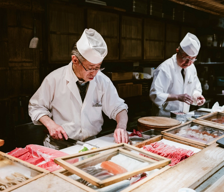 Two sushi chefs in traditional white uniforms preparing fresh sashimi behind a counter filled with trays of raw fish in a Japanese restaurant kitchen