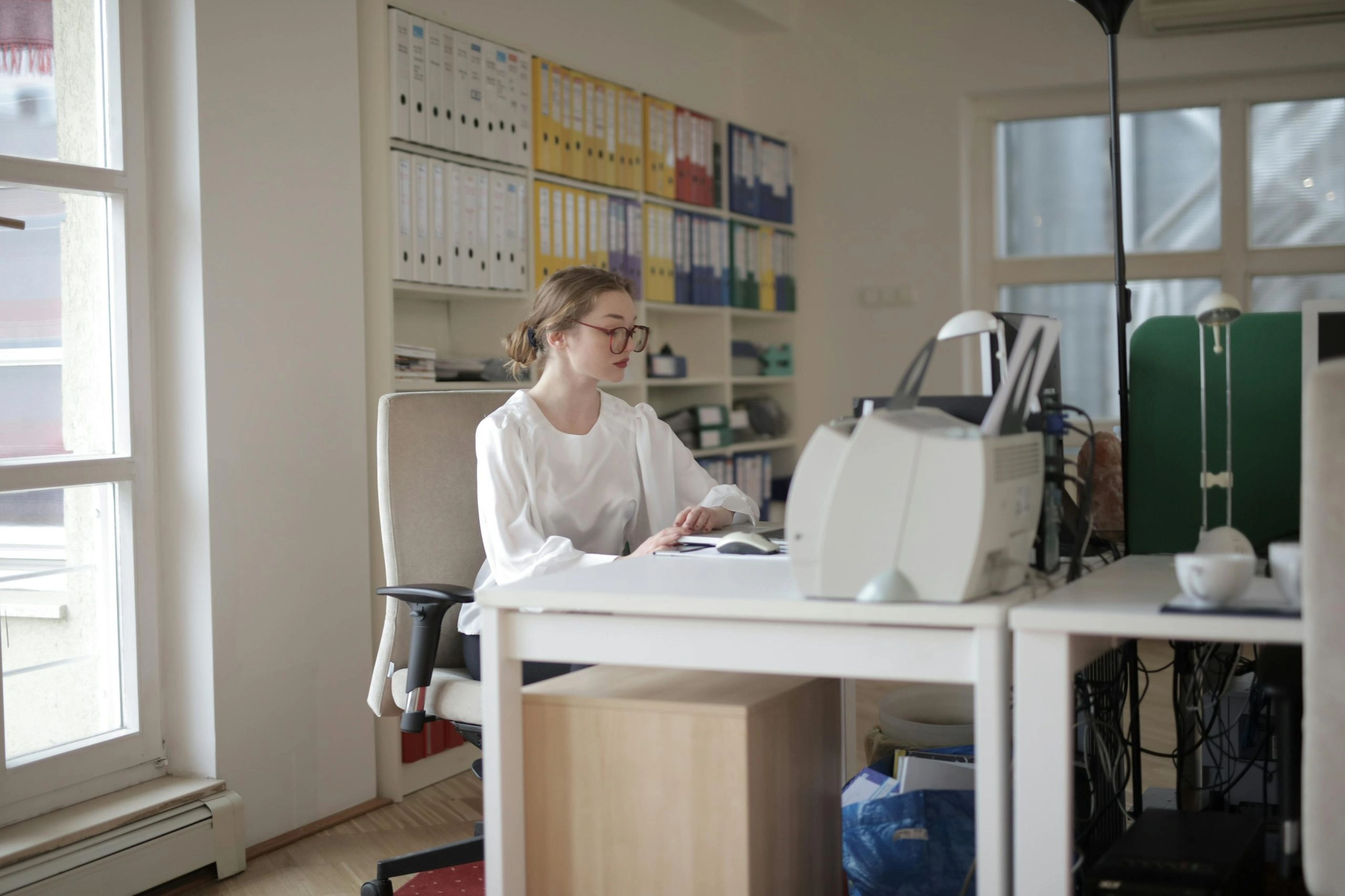 Young woman with brown hair tied back, wearing glasses and a long sleeved white shirt sitting at computer to illustrate article about bookkeeping 101