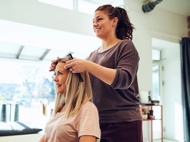 Hairdresser smiling while styling a client’s hair in a bright, modern salon with large windows and hair products on display
