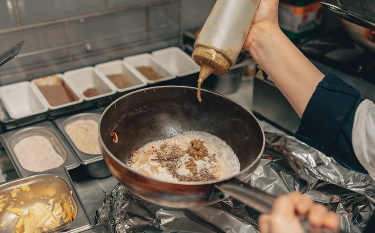 Close-up of a cook adding sauce to a pan with ingredients in a kitchen.